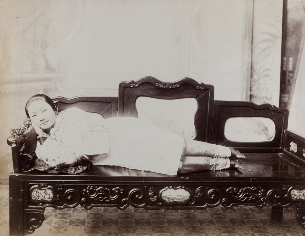 A woman with bound feet, lying on a bench in a photographer’s studio, with a book