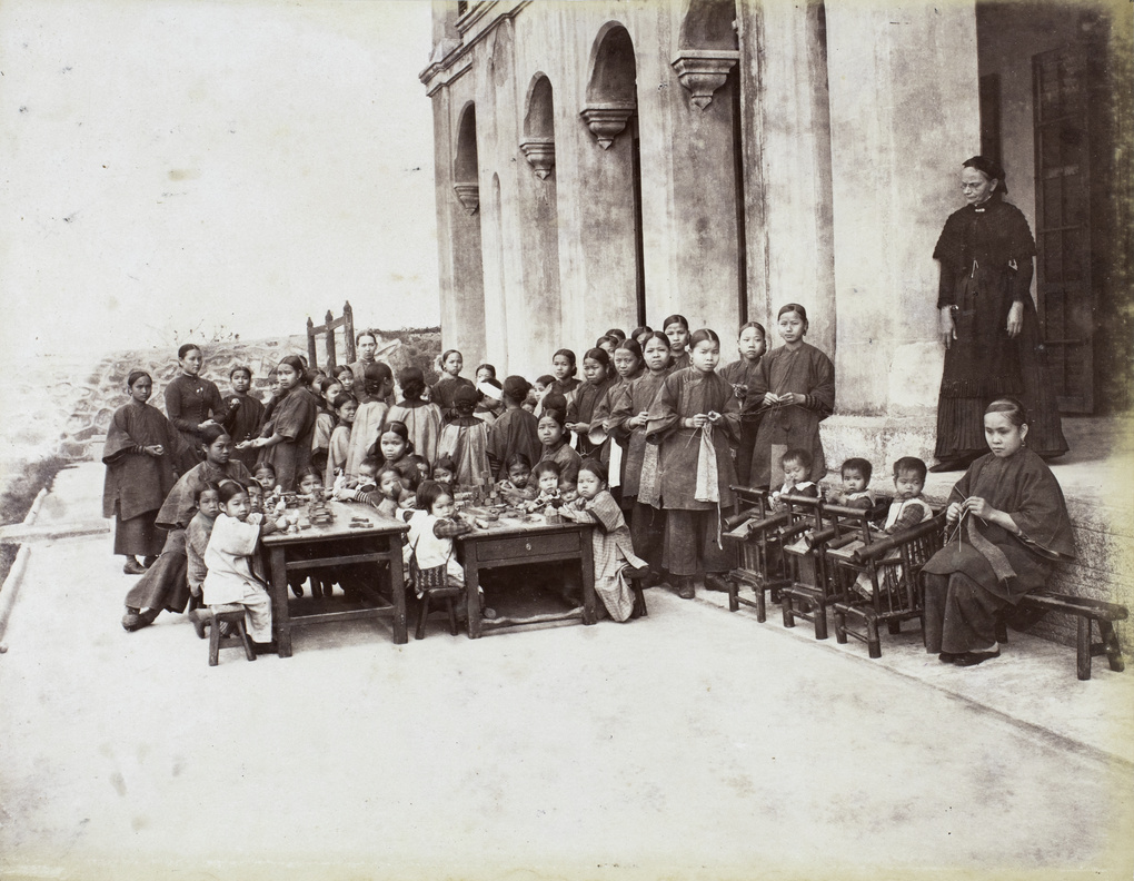 A large group of girls, probably under missionary care, eating, knitting and standing next to pillars of a Church