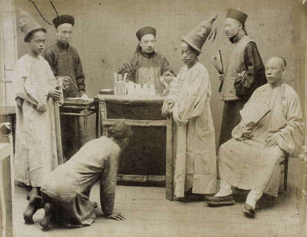A man kneeling in front of the magistrate, with others wearing hats and carrying fans