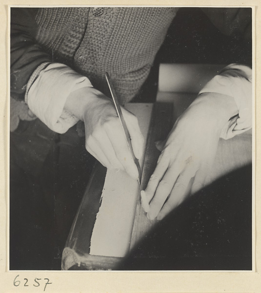 Interior of a scroll-mounting shop showing a man trimming the edge of a scroll painting with a steel paper knife