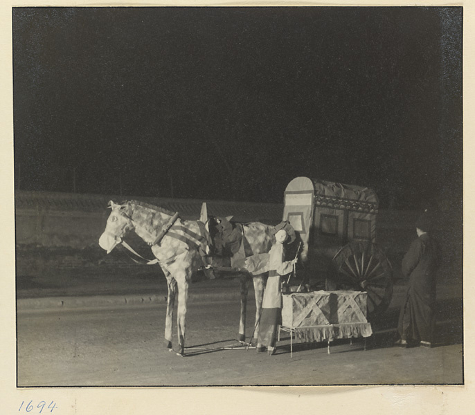 Man at night standing next to paper funeral objects including horse, cart, and human figure