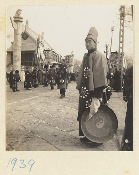 Musician holding a gong with yin-yang symbol and men with umbrellas, staffs, and banners in front of building with hua biao awaiting the start of a funeral procession