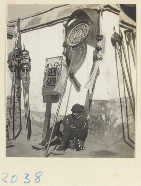 Member of a wedding procession seated by a wall with lanterns, flags, inscribed banners, draped mirrors, and staffs