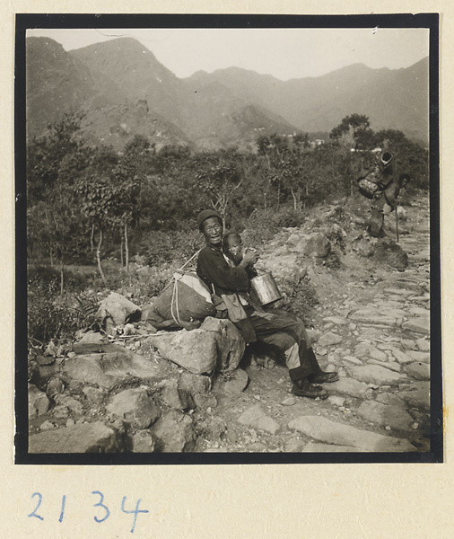Seated pilgrim with backpack holding child and metal bucket on Miaofeng Mountain