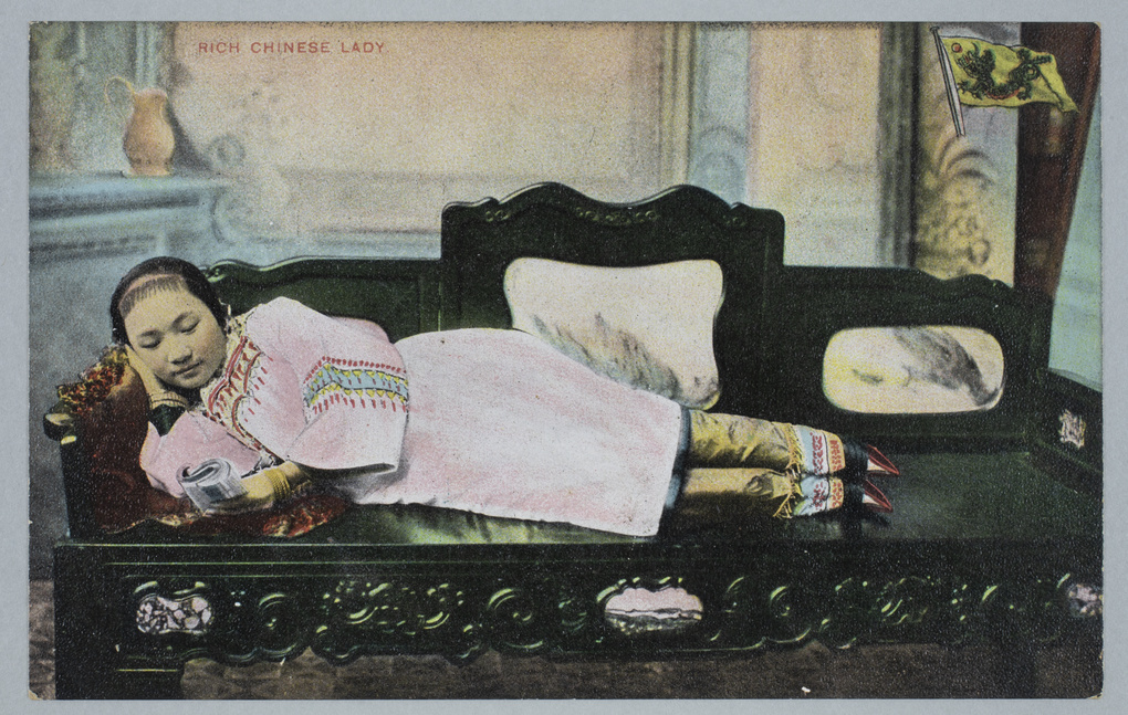 A woman with bound feet, lying on a bench in a photographer’s studio, reading a book