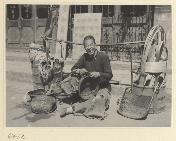 Mat mender with tools and reeds repairing a basket with signs in background