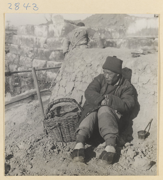 Ice-cutter hauling a block of ice past a man sitting with a basket