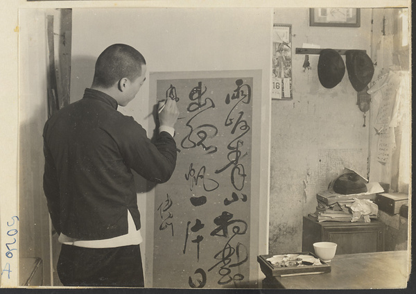 Interior of a scroll-mounting shop showing a man retouching a scroll hanging on a drying board called a quan