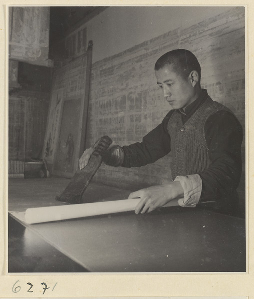 Interior of a scroll-mounting shop showing a man adhering a backing to a scroll painting by tapping with a stiff brush called a zhong pi shua