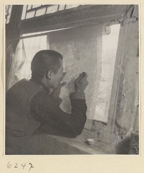 Interior of a scroll-mounting shop showing a man at a window repairing the back of a scroll painting