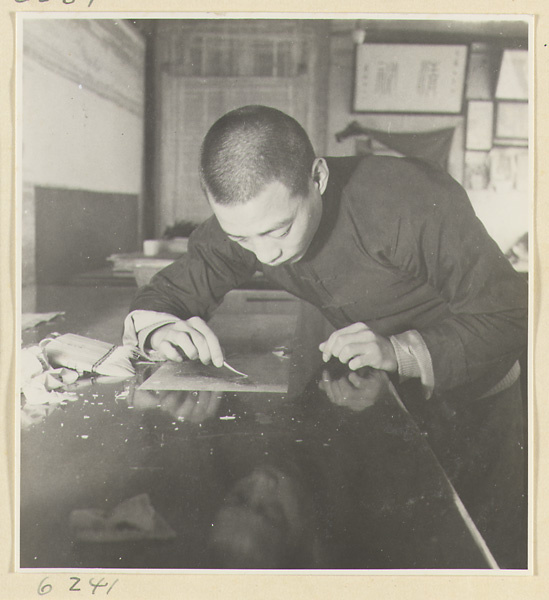 Interior of a scroll-mounting shop showing a man using iron tweezers to remove bits of paper from the back of a scroll painting
