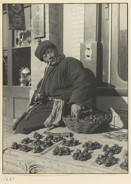 Street vendor smoking a pipe and selling dried jujubes next to a brass-workers shop with a sign offering all kinds of ink pots