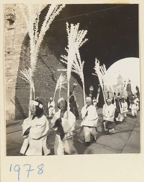Children dressed in white carrying paper snow willows and banners and passing through a city gateway in a funeral procession
