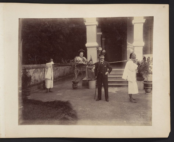 Unidentified man standing in front of woman sitting in sedan chair with two Chinese bearers, Canton