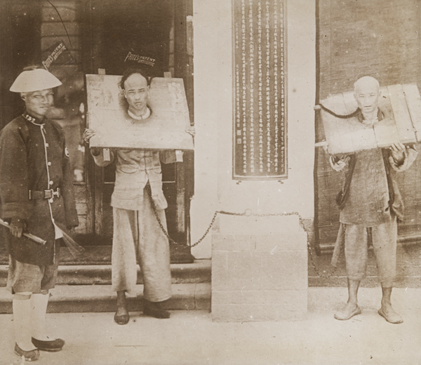 Two prisoners in cangues, with a Chinese policeman, chained outside Watson's Pharmacy, Hong Kong