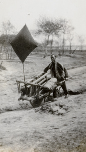 A porter with a loaded wheelbarrow assisted by a sail, North China