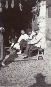Three men sitting over a threshold, and a man in the shadows holding a pipe
