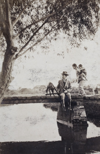 Tom Hutchinson sitting on a bridge with a hunting dog and rifle while a woman carrying a child on her back is crossing, Shanghai
