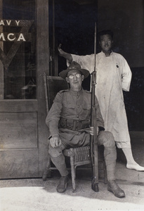 Hostel worker standing behind Floyd Tangier Smith, an American Company Shanghai Volunteer Corps member on duty at the entrance to the Navy YMCA, Shanghai, 1925