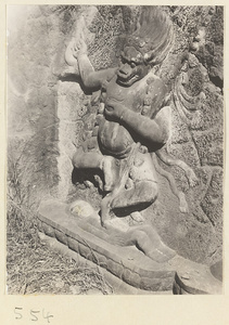 Animal-headed relief figure wearing necklace of skulls, carrying a body, and standing on a prone figure carved into the hillside at Yuquan Hill