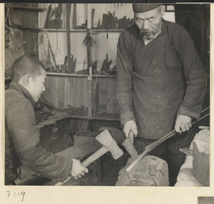 Blacksmith and assistant hammering a piece of heated metal in a shop