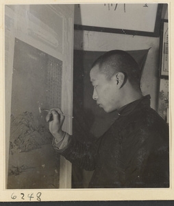 Interior of a scroll-mounting shop showing a man touching up areas of loss on a scroll painting as it hangs on a drying board called a zhuang ban