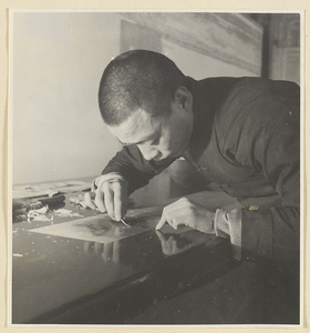 Interior of a scroll-mounting shop showing a man using iron tweezers to remove bits of paper from the back of a scroll painting