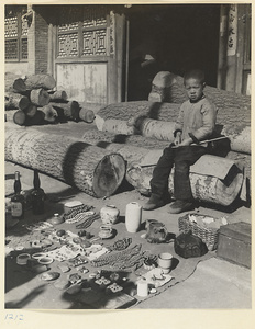 Boy sitting on a pile of logs and hawking jewelry and decorative objects outside a shop with door couplets