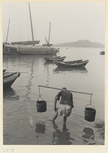 Man with buckets hung from a shoulder pole wading next to fishing boats at anchor on the Shandong coast