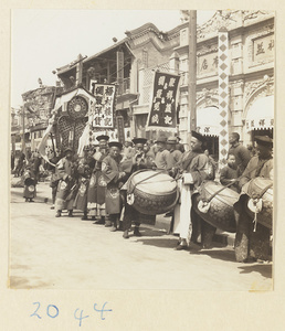 Members of a wedding procession standing in front of shops holding fan-shaped screen and draped mirror and playing musical instruments
