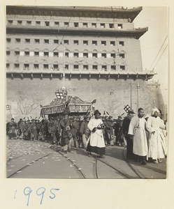 Principal mourner wearing white cap, man with drum keeping a beat for coffin bearers, and draped coffin being carried in front of city gate