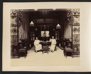 Two young Chinese men sitting at table in room with carved wood furniture and partitions, Canton