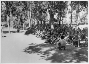 Mass of soldiers sitting in the shade, listening to a speaker