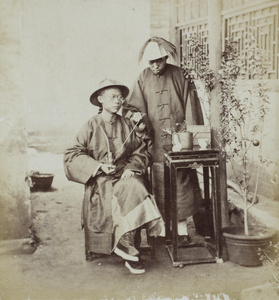 A Manchu man and a servant, posed with a tobacco pipe, table, teapot, and fruit tree, Beijing