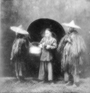 Studio photograph of two men wearing straw rain coats, with a man holding an umbrella and a basket