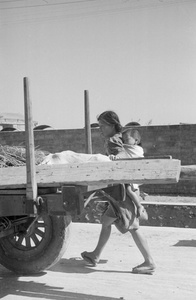 Woman carrying a child on her back, walking beside a cart
