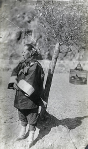 A woman with bound feet beside a bird cage hanging in a tree