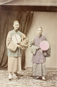 Two men with fans in a photographer’s studio, with posing stands and a backdrop curtain